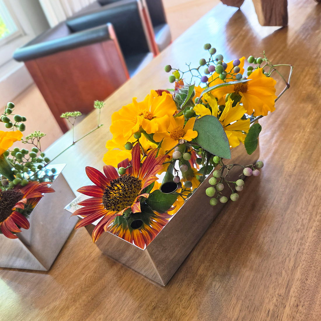 Polished stainless steel 8-inch rectangular Ikebana vase holding vibrant yellow and orange sunflowers on a wooden table.